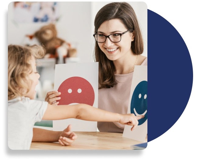 A smiling teacher holding two sheets of paper, one with a sad face and one with a happy face. A child is pointing at the smiling, happy face.