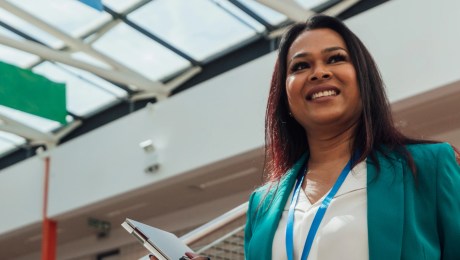 A medium close-up portrait of a female principal stood confidently on the stairs into the school hall. She is carrying a laptop in one of her hands.