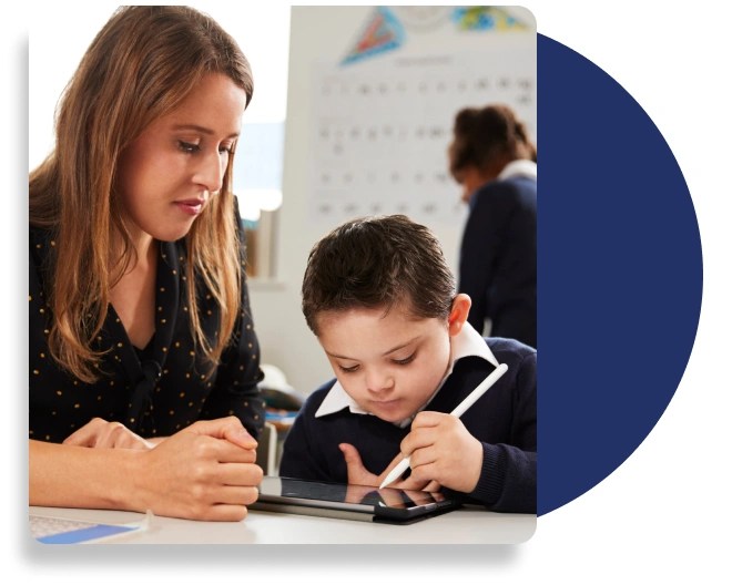 A teacher and a primary school aged student sitting at a desk. The learner is using a stylus to write on a tablet computer.